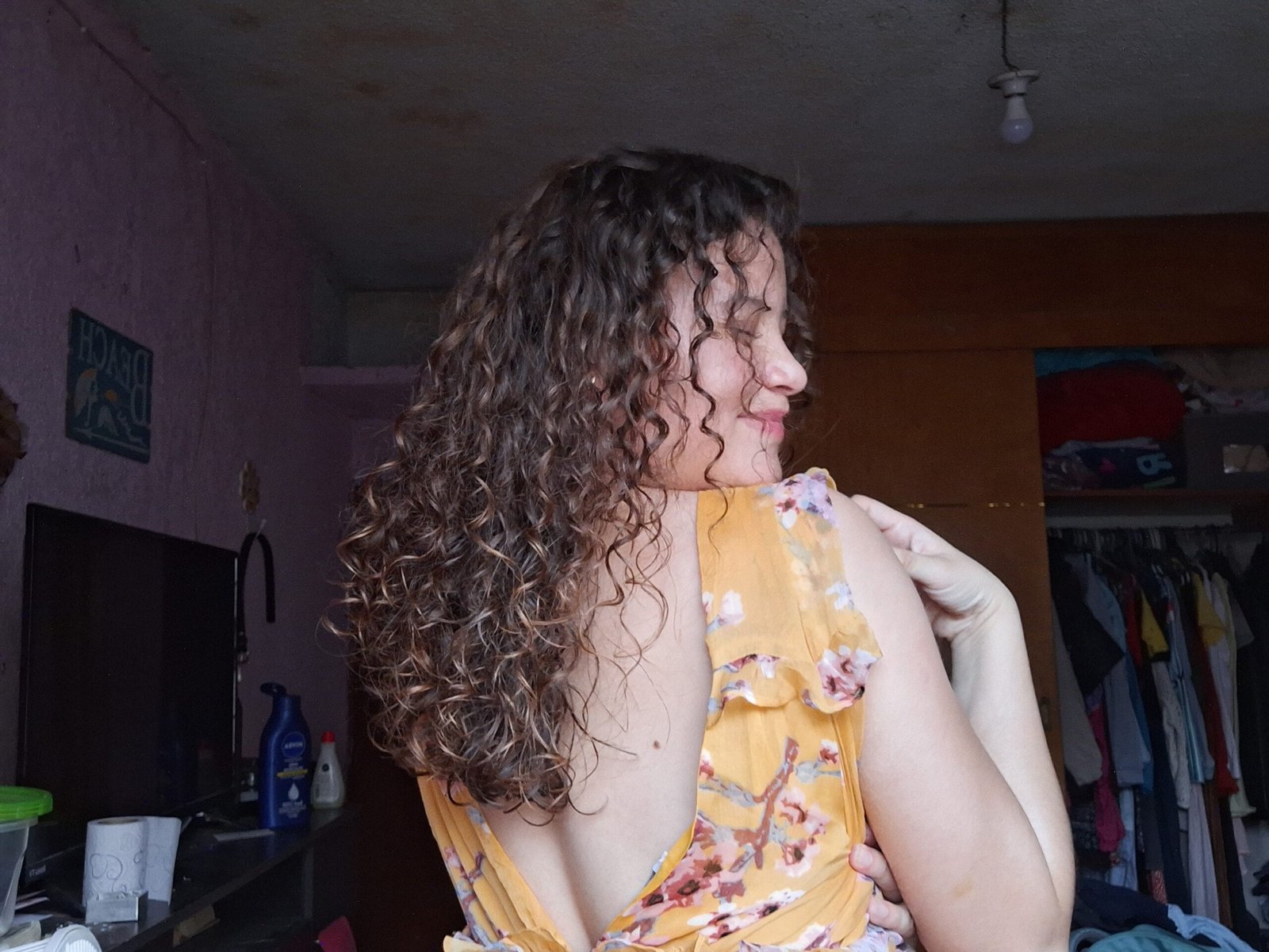 mujer de espaldas, rizada 3a, castaña clara, con chinos definidos e hidratados mediante mascarilla casera y fácil, cabello un poco largo, blanca, sonriente, cabello voluminoso y rizos compactos, con vestido amarillo