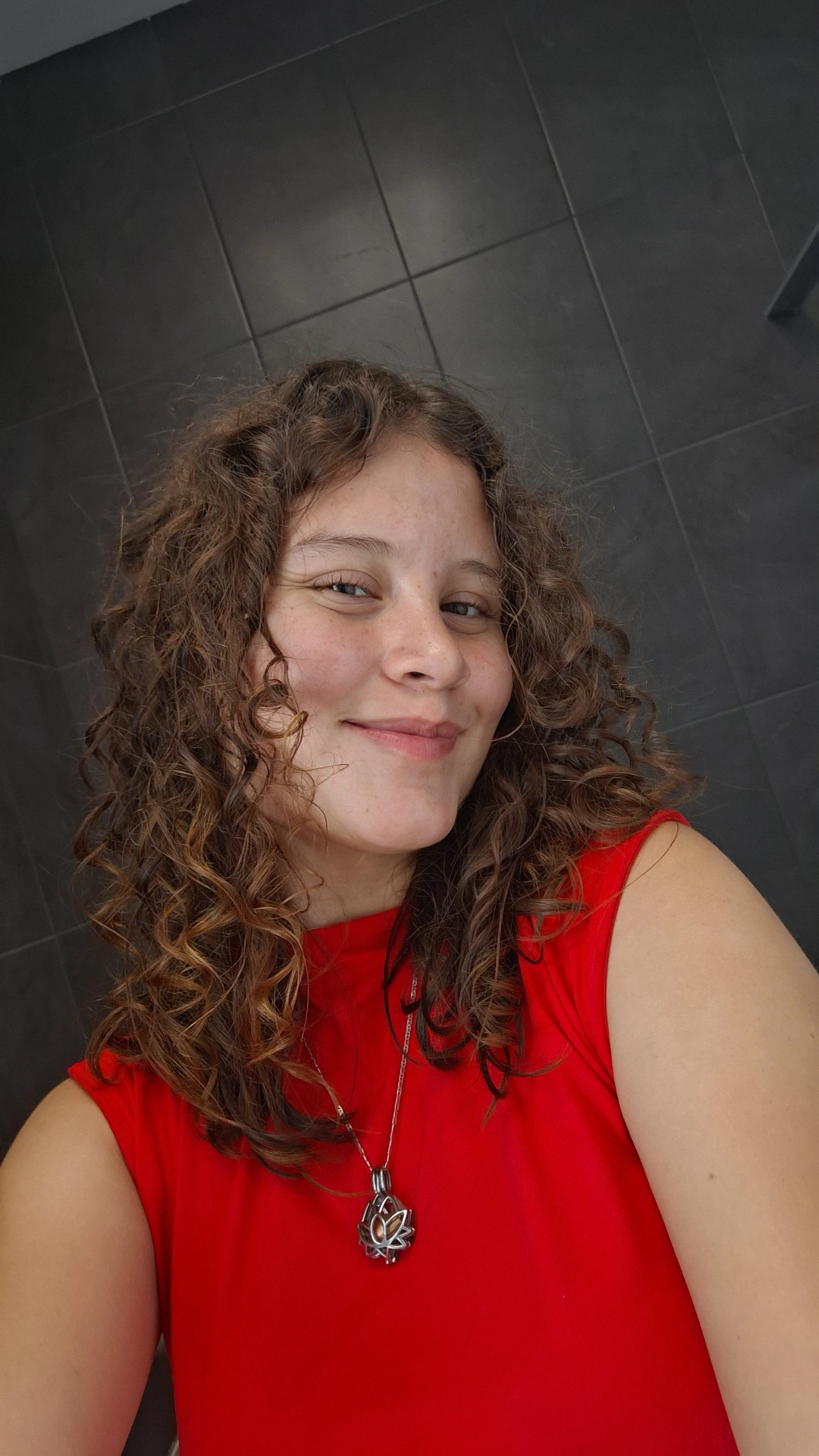 mujer con cabello rizado tipo 3a, sonriente, blanca, con cabello poco voluminoso, castaña clara, blusa roja, cejas poco pobladas, ojos chiquitos, un collar largo, cabello con rizos poco definidos y cabello a la altura de los hombros, previamente a aplicarse una mascarilla hidratante, con rizos sin brillo