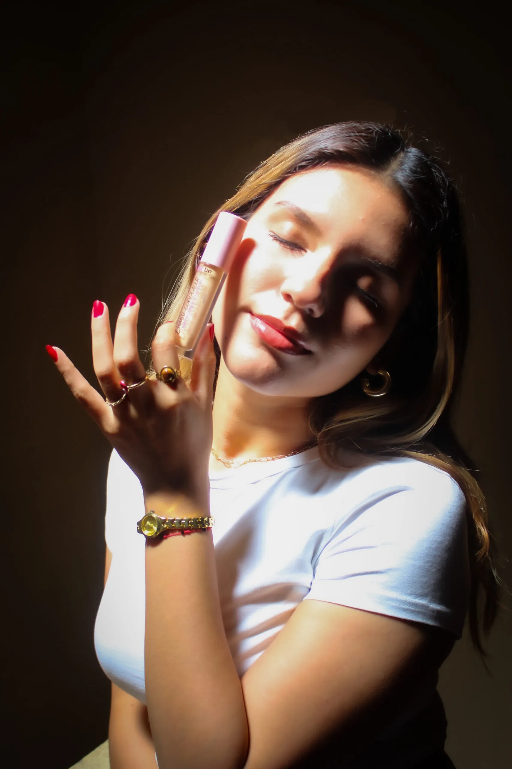 Mujer rubia con joyería dorada blusa blanca con uñas rojas, con ojos cerrados, de fondo oscuro liso, ojos cerrados, sosteniendo un corrector como producto de maquillaje con su mano izquierda para su rutina de maquillaje, con rostro relajado.