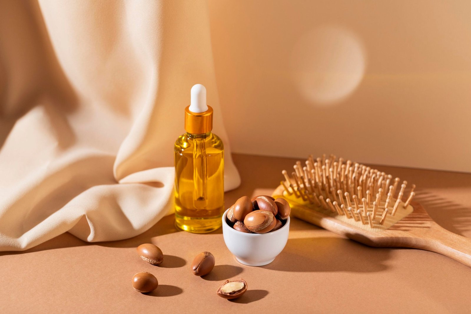 gotero con aceite de argán para el cabello, junto a una pequeña taza llena de semillas de argán y a lado un cepillo de cabello y fondo con colores neutros