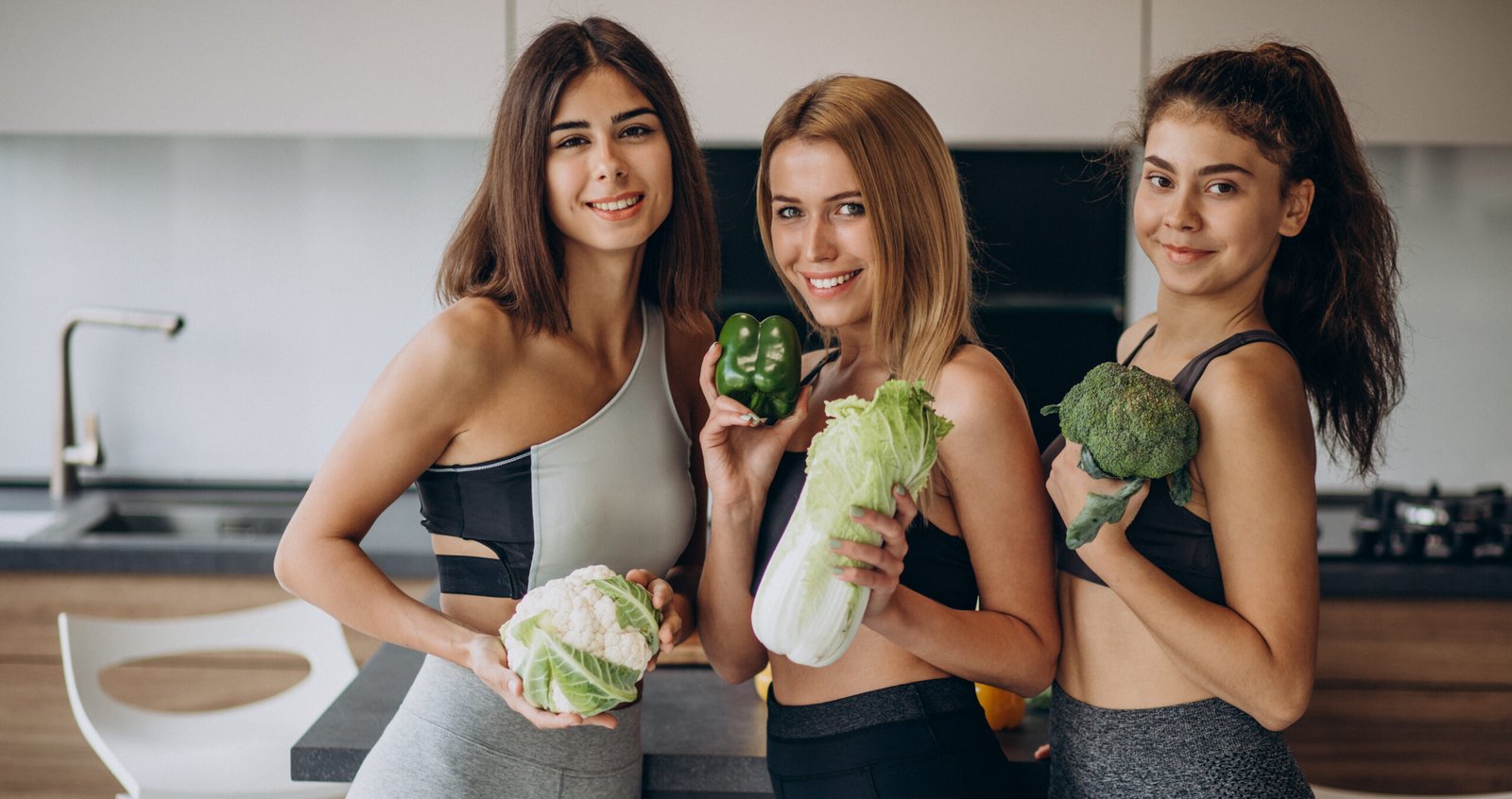 Tres mujeres en ropa deportiva sonríen mientras sostienen vegetales frescos, promoviendo un estilo de vida saludable con ejercicio fácil en casa y una buena digestión.