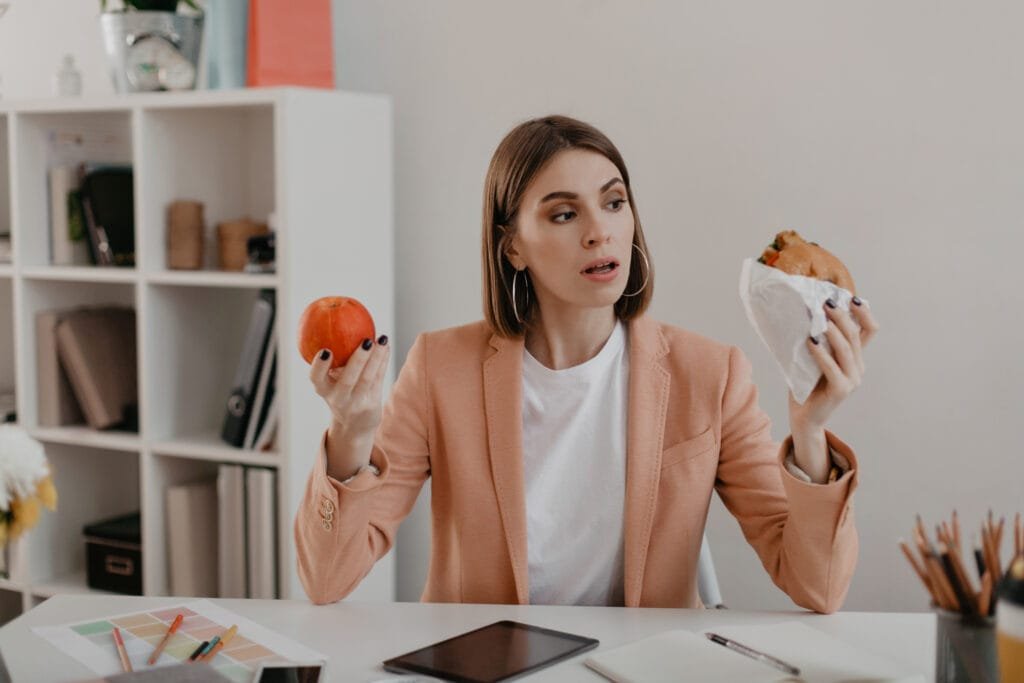 mujer joven trabajando en una oficina con cabello corto, piel saludable y suave, sin texturas y poco maquillaje decidiendo entre dos alimentos , un alimento sano y otro no saludable.