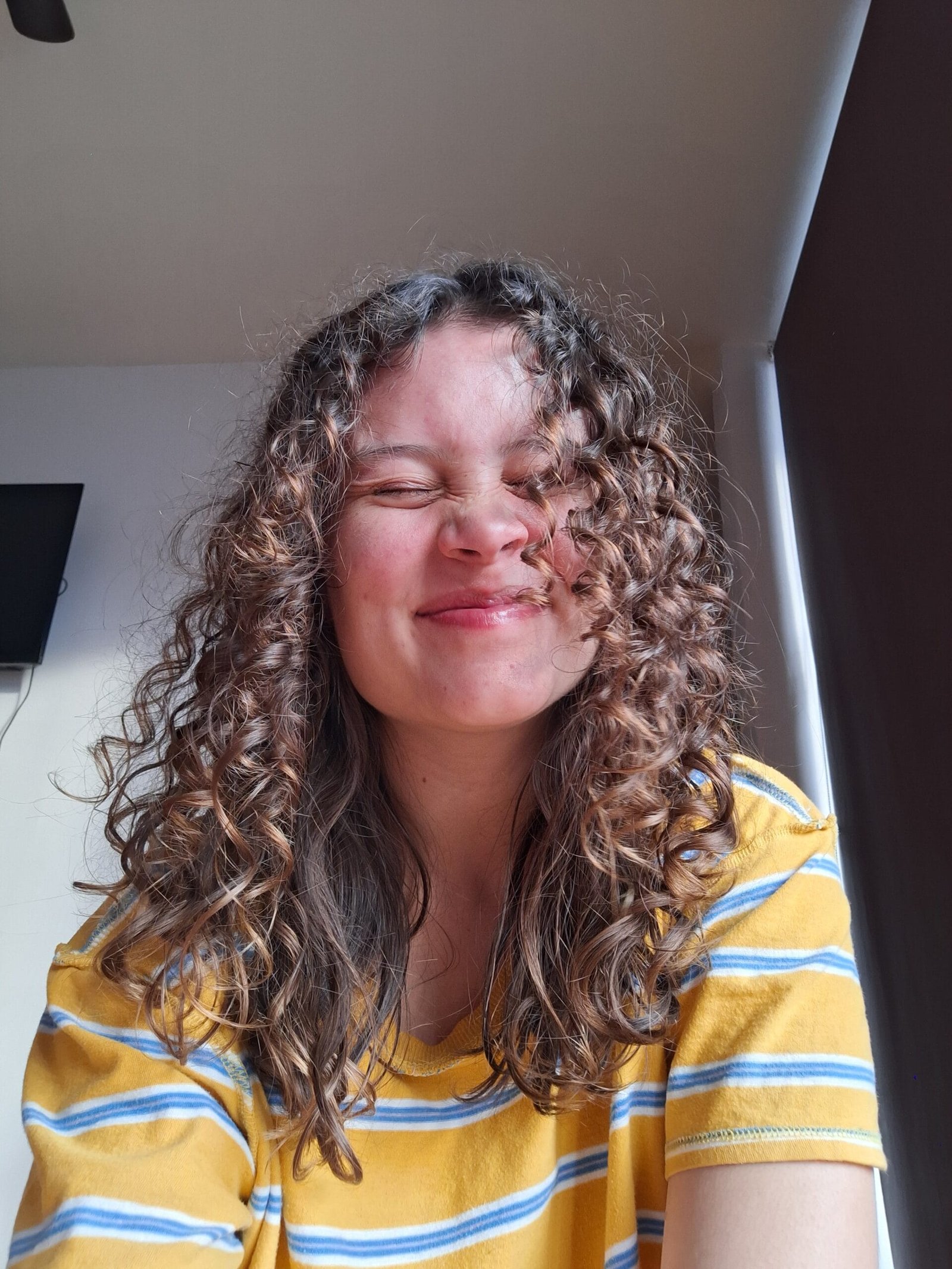 mujer de rizada 3a, castaña clara con destellos amarillos, playera amarilla rayada, cuero cabelludo dividido en dos, blanca, sonriente, con ojos cerrados, cejas poco pobladas, nariz y ceño fruncidos, con fondo blanco