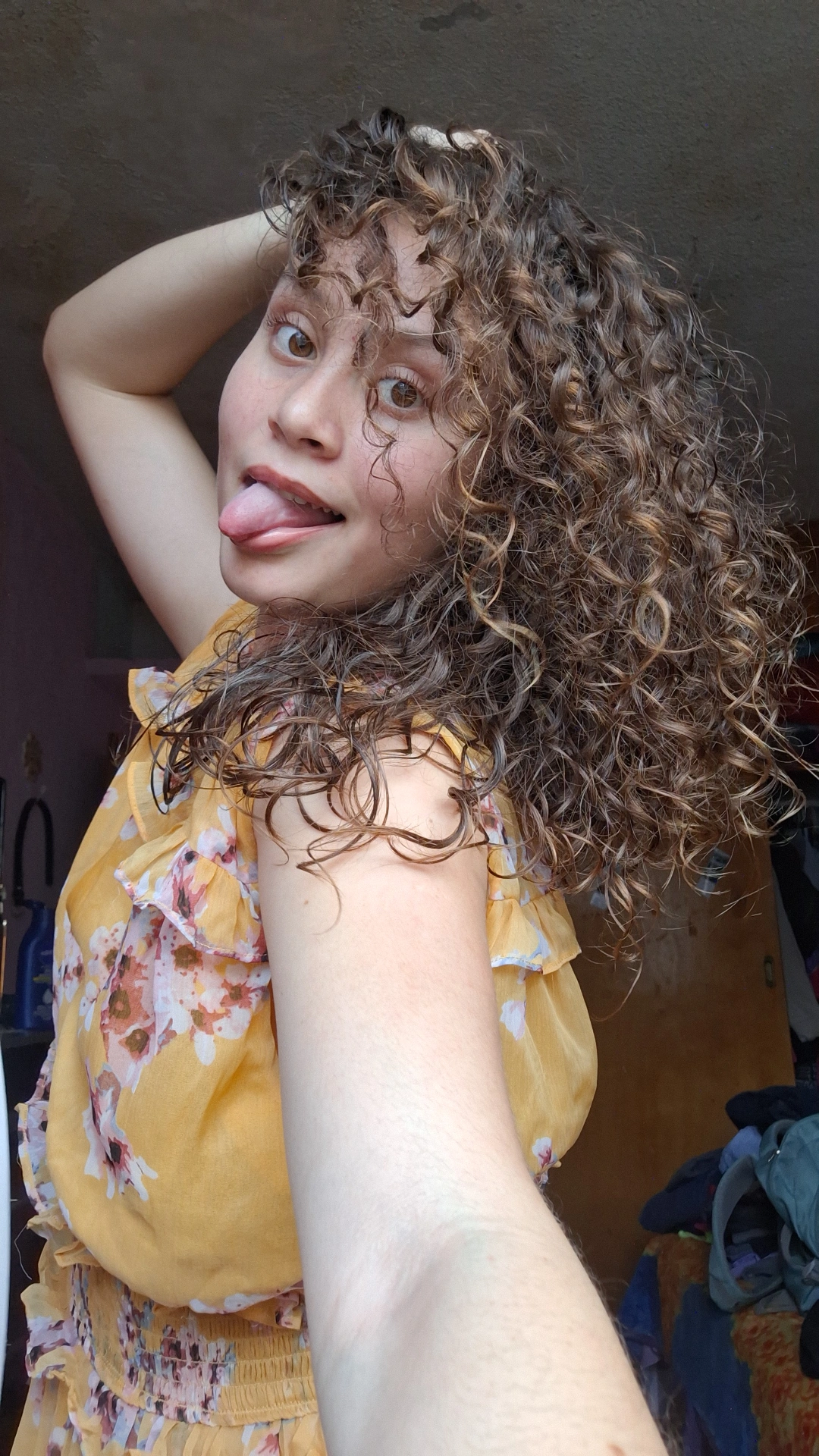 mujer con cabello muy chino, con rizos 3a, cabello castaño muy claro, despeinada y natural, peinada con difusor, con un vestido amarillo, blanca, con lengua de fuera junto con gestos propios y alocados, cabello voluminoso, definido con un difusor, mujer bonita enseñando sus cualidades