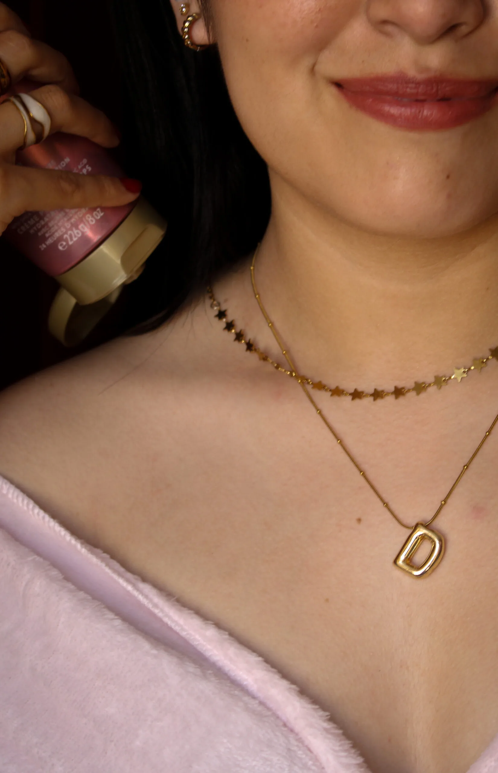 mujer milenial  con lunares, collares dorados en el cuello, labial rosa, ligeramente destapada de los hombros aplicándose crema para su tipo de piel seca y sensible 