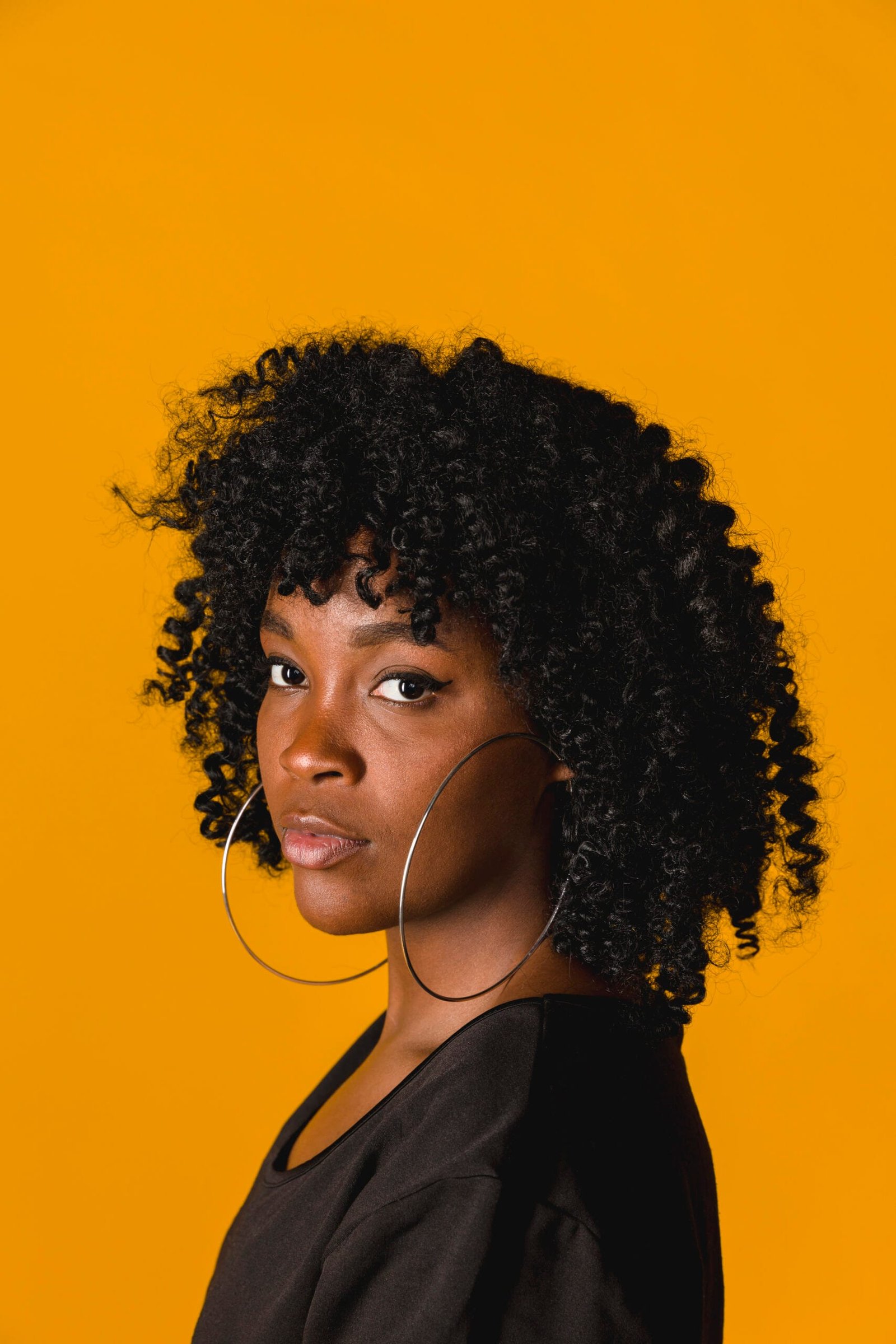 mujer afro 4a, rizos un poco cerrados y definidos, volumen medio, de tez morena, cabello negro, semblante serio, labios gruesos, aretes grandes, playera negra, maquillada y fondo amarillo