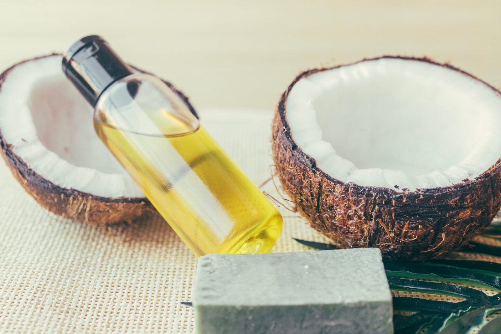 frasco con aceite de coco que ayuda con crecimiento del cabello, colocado a su lado se encuentra dos mitades de un coco, un jabón y una palma de coco