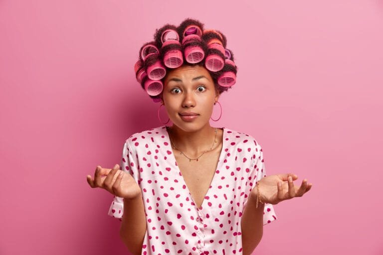mujer con tubos para darle más forma a su cabello afro, dejando una textura más chino, estando confundida o indecisa, con cejas levantadas, ojos abiertos, tez oscura, sonrisa chica, aretes grandes, manos en forma de confusión, playera estampada con un collar en el cuello, generando duda y desconcierto