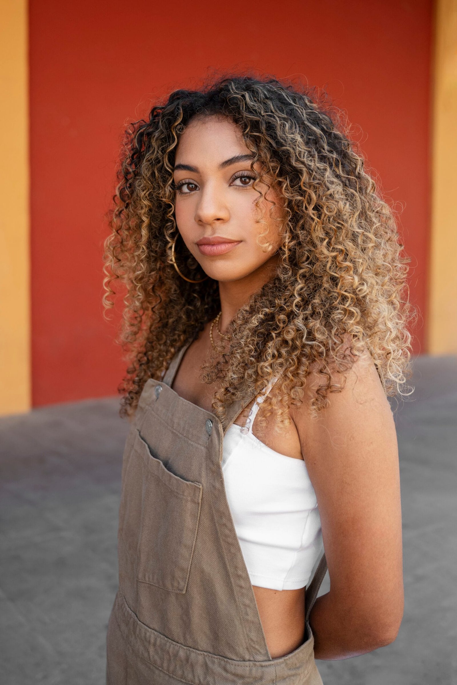 mujer con cabello chino tipo 3c, mediano con gran volumen, pintado y difuminado de dos colores, en capas, con rizos definidos, un poco despeinada, con cabello partido en dos, cejas marcadas, labios gruesos, nariz fina, vestida con un overol y un top debajo 
