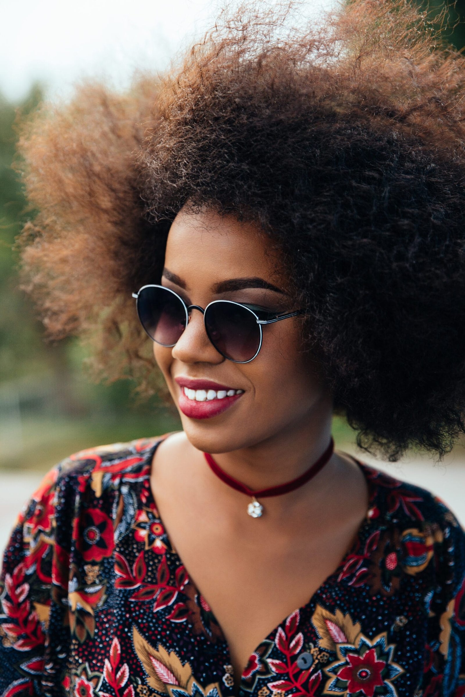 mujer afro con rizos 4b, cabello despeinado, desarreglado, café, con rizos semi cerrados, cabello corto y esponjado. sonriente, cejas marcadas y definidas, con lentes de sol, una blusa colorida y floreada y un collar
